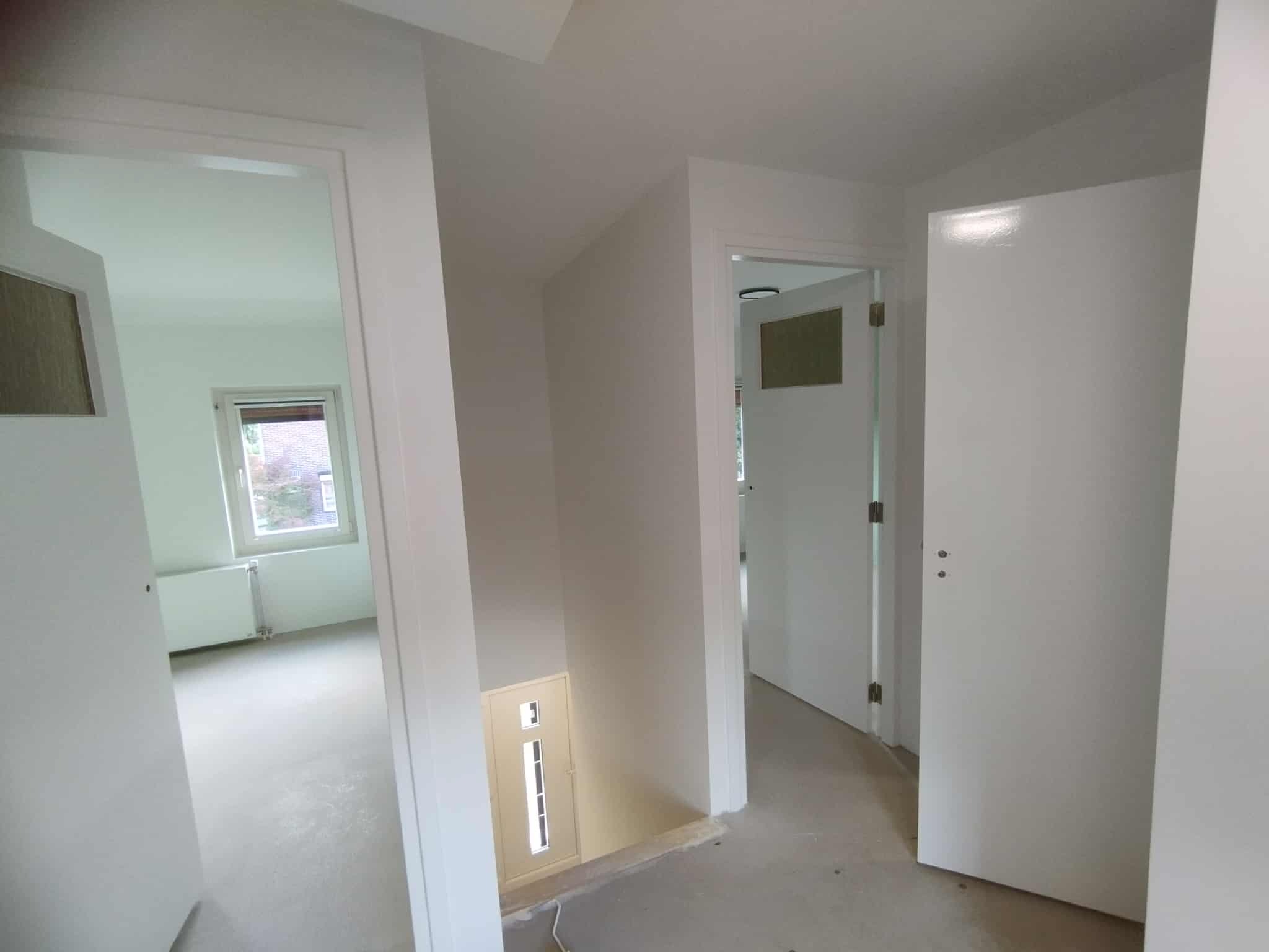 Białe, świeżo pomalowane wnętrze mieszkania z otwartymi drzwiami, widok na pokój z oknem. Podłoga w trakcie prac wykończeniowych.