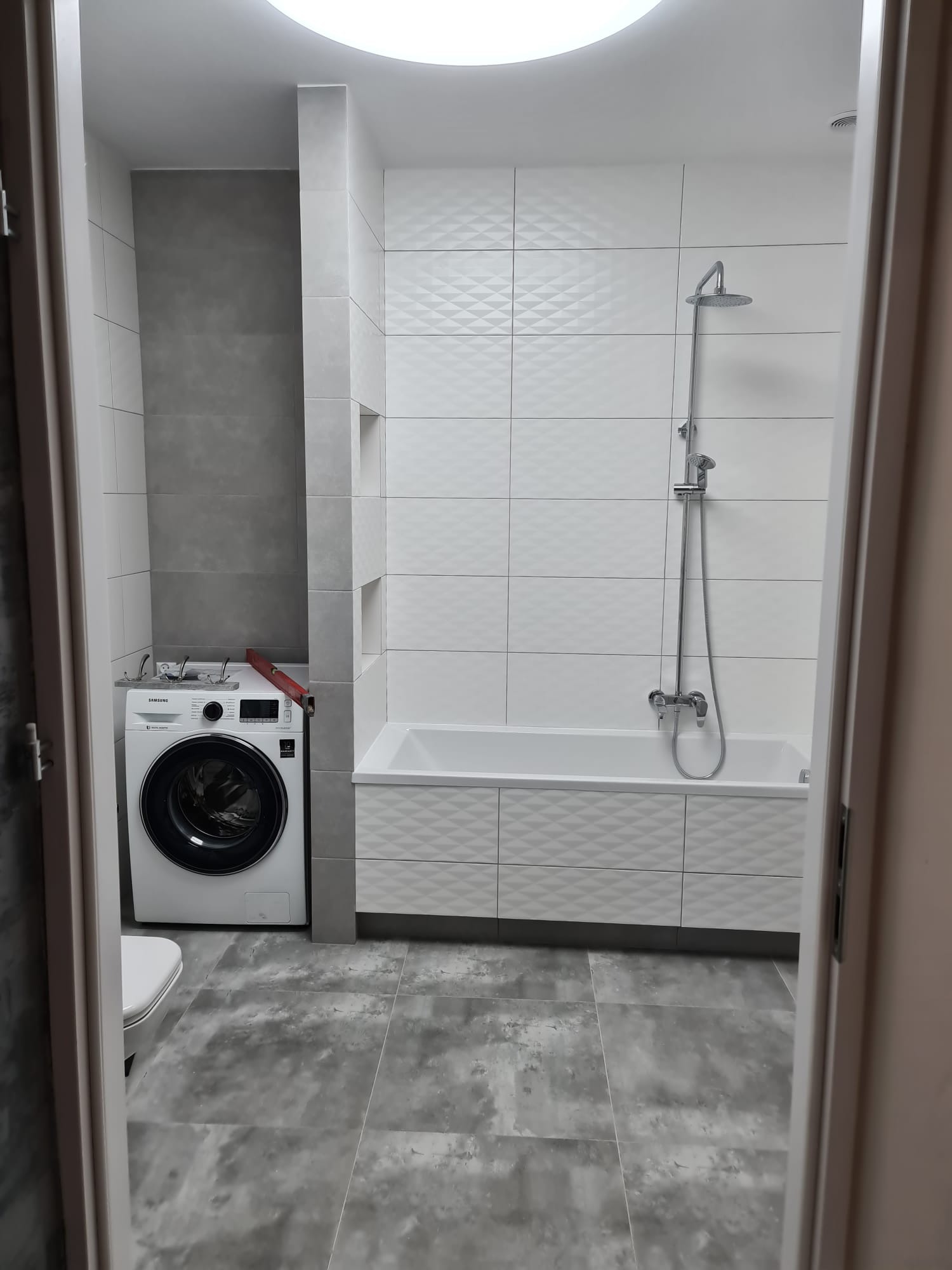 Wykończona łazienka w szaro-białej tonacji. Widoczna pralka, wanna z zabudową i armatura. Płytki na ścianach z subtelnym wzorem. Podłoga wyłożona szarymi płytkami.