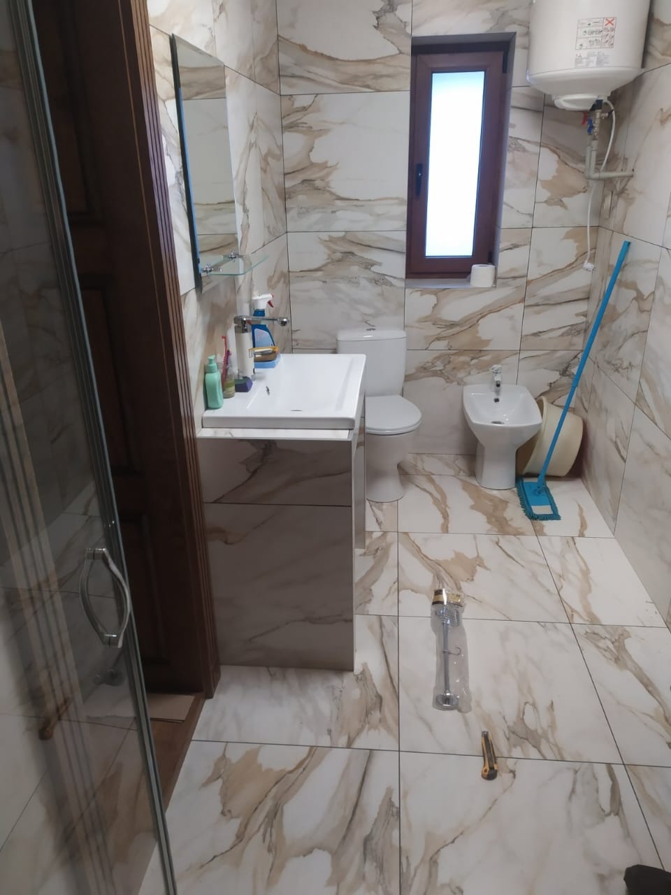 Wykończona łazienka z marmurowymi płytkami, białą armaturą i drewnianymi akcentami. Widoczny fragment kabiny prysznicowej i akcesoria montażowe na podłodze.