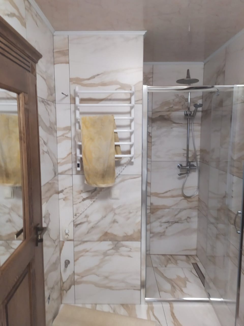 Wykończona łazienka z kabiną prysznicową i grzejnikiem drabinkowym, płytki imitujące marmur na ścianach i podłodze, drewniane drzwi po lewej stronie.