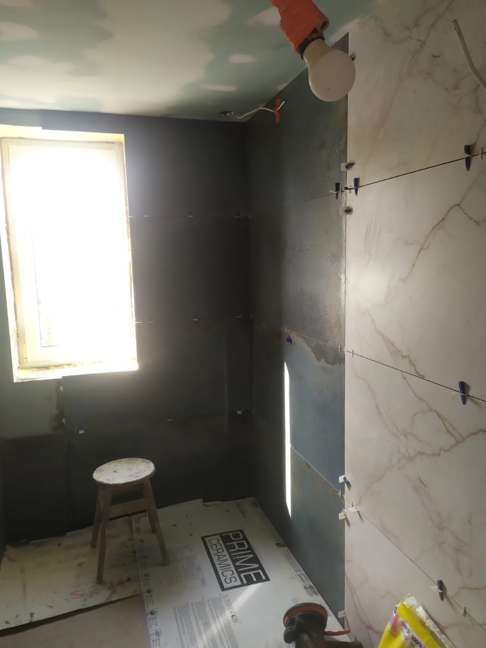 Wnętrze łazienki w trakcie prac glazurniczych: częściowo wyłożone płytki imitujące marmur, ciemne płytki na ścianach, widoczne krzyżyki dystansowe i narzędzia.