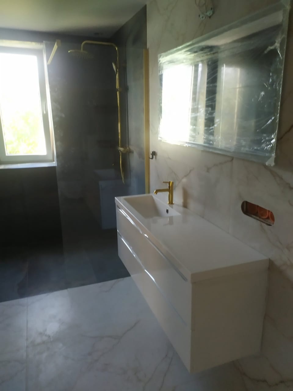 Nowoczesna łazienka z marmurowymi płytkami, białą szafką pod umywalką i złotą armaturą. Lustro zabezpieczone folią. Widoczny fragment kabiny prysznicowej.