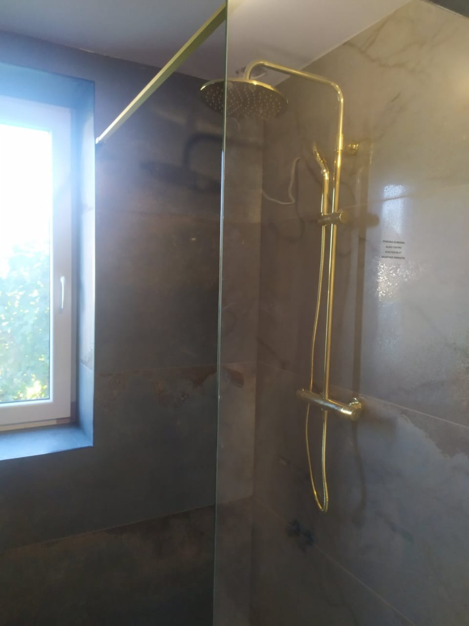 Nowoczesna kabina prysznicowa z deszczownicą w odcieniach złota, na tle płytek imitujących kamień. Widoczne okno z białymi ramami.