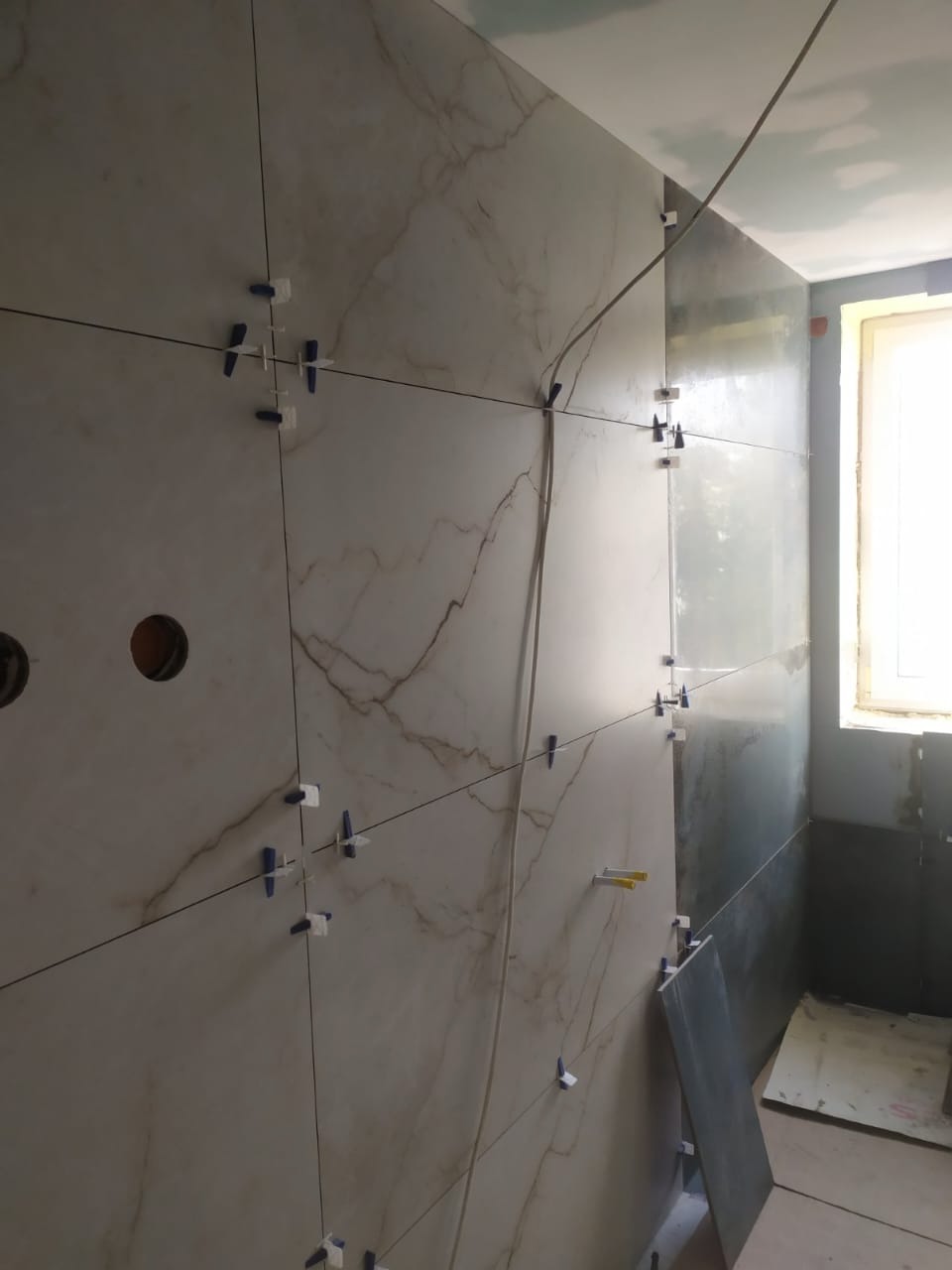 Ściana w trakcie układania płytek imitujących marmur, z użyciem klipsów poziomujących. Widoczne otwory na instalacje i fragment okna.