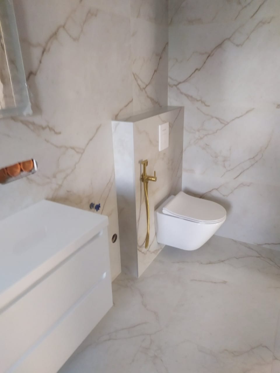 Wykończona łazienka z marmurowymi płytkami, podwieszaną toaletą z złotą słuchawką bidetową i białą szafką pod umywalkę. Minimalistyczny design.