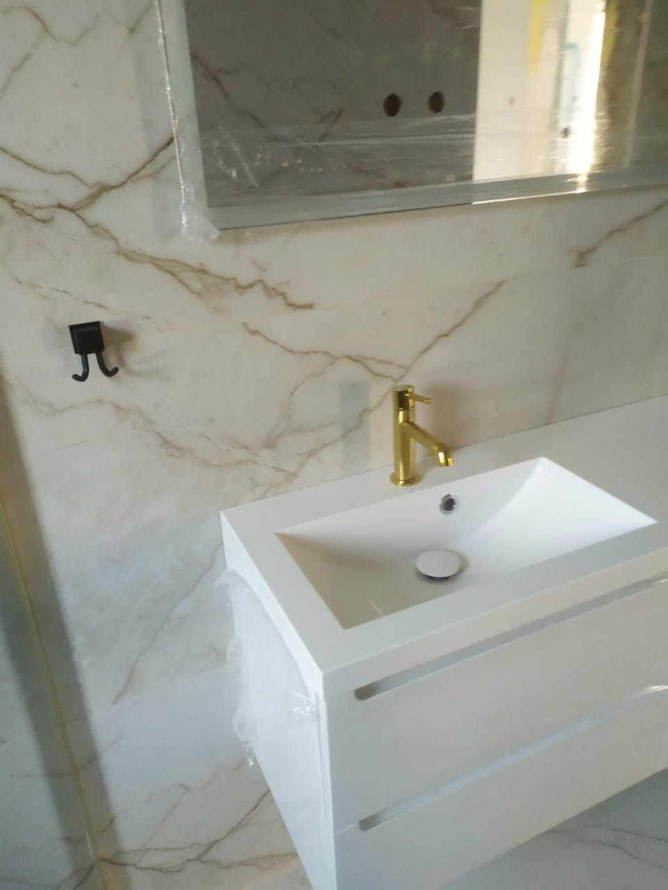 Biała szafka pod umywalkę z złotą baterią na tle marmurowej ściany. Nowoczesna łazienka z lustrem w folii ochronnej i czarnym wieszakiem na ręczniki.