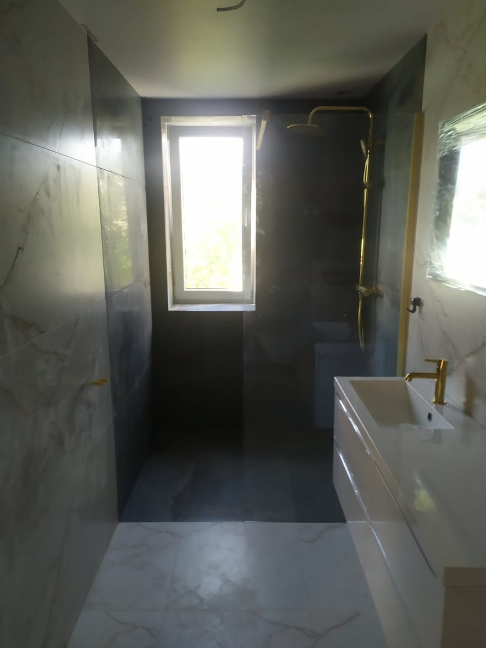 Nowoczesna łazienka z marmurowymi płytkami, złotą armaturą i kabiną prysznicową typu walk-in. Okno doświetla wnętrze. Minimalistyczny design.