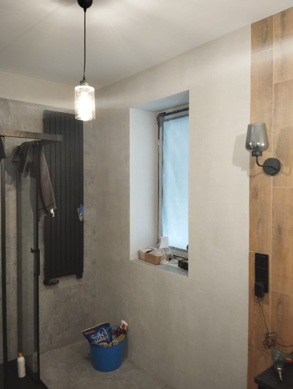 Wykończona łazienka z szarą kabiną prysznicową, grzejnikiem drabinkowym i oknem. Na ścianie drewnopodobne płytki i lampa. Na podłodze stoi niebieskie wiadro z chemią.
