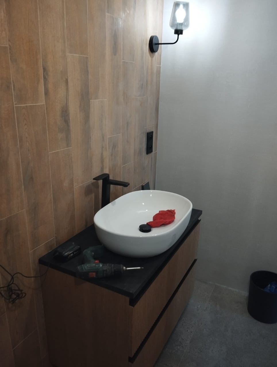 Nowoczesna łazienka z drewnopodobnymi płytkami, białą umywalką nablatową i czarną armaturą. Widoczny czarny blat, narzędzia i oświetlenie.