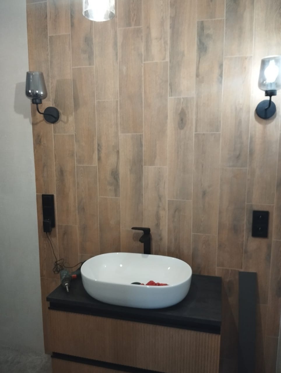 Ukończona łazienka: owalna umywalka nablatowa, czarna bateria, drewnopodobne płytki ścienne, czarne kinkiety. Widoczny fragment wiertarki.