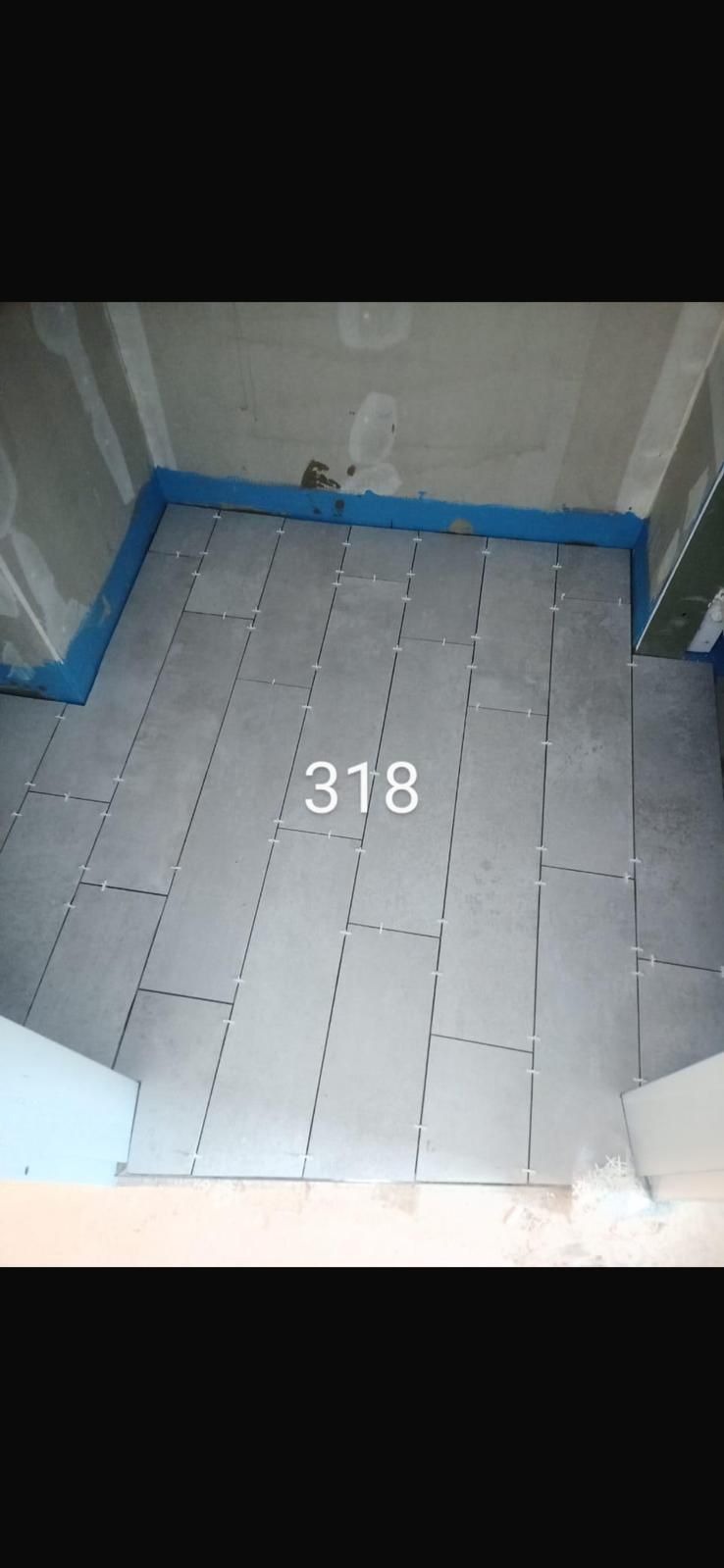 Układanie szarych płytek podłogowych w łazience, widoczne krzyżyki dystansowe i niebieska taśma uszczelniająca przy ścianach. Etap prac wykończeniowych.