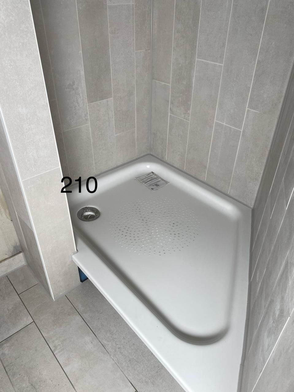 Narożny brodzik z odpływem w kabinie prysznicowej wyłożonej szarymi, pionowymi płytkami. Minimalistyczny design, jasne wnętrze łazienki.