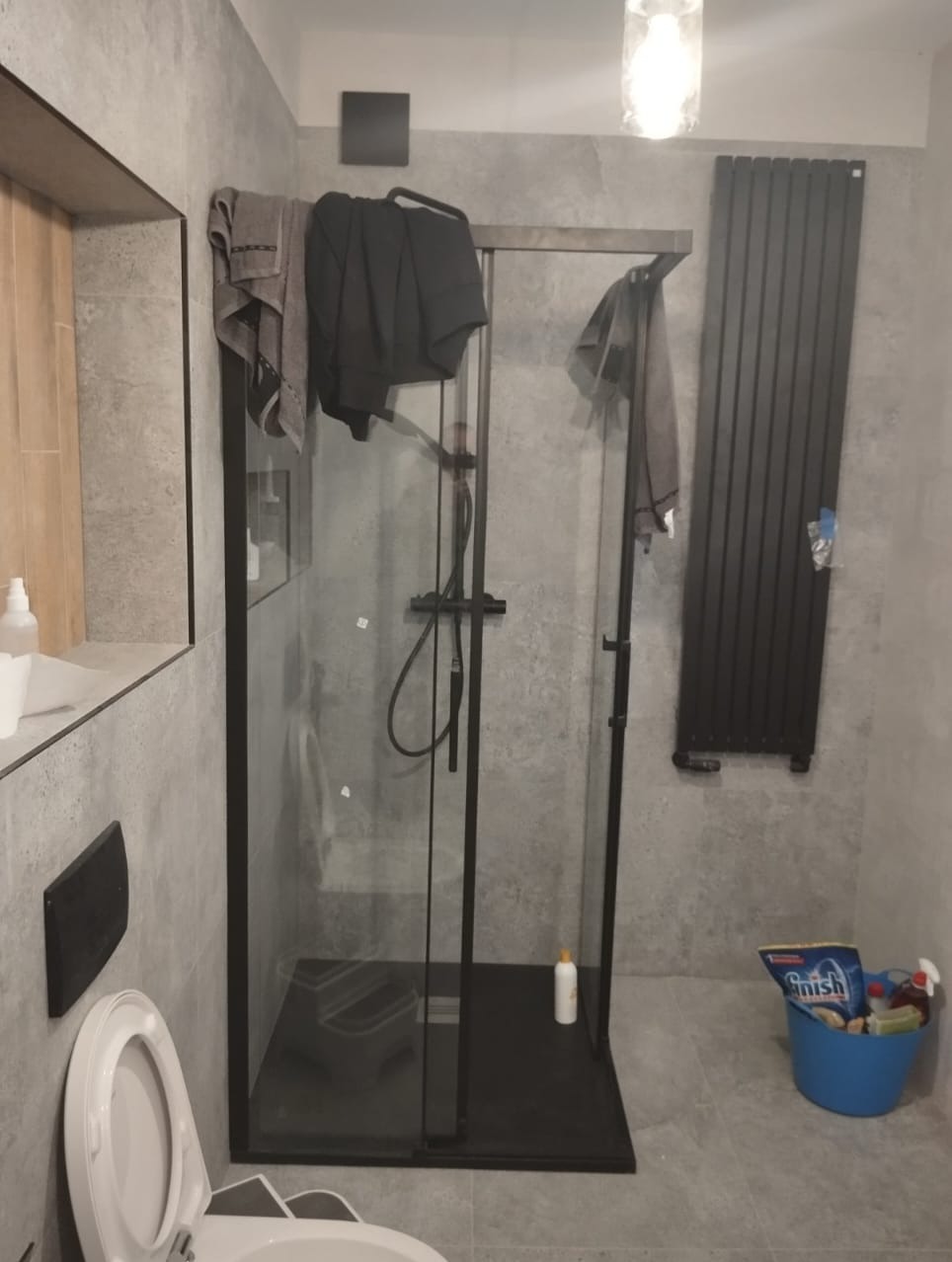 Nowoczesna łazienka z kabiną prysznicową typu walk-in, szarą armaturą i płytkami imitującymi beton. Widoczny fragment toalety i grzejnik drabinkowy.