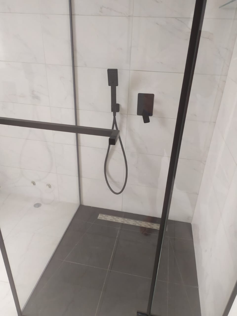 Nowoczesna kabina prysznicowa z czarną armaturą i odpływem liniowym. Białe marmurowe płytki na ścianach, szare płytki podłogowe. Minimalistyczny design.