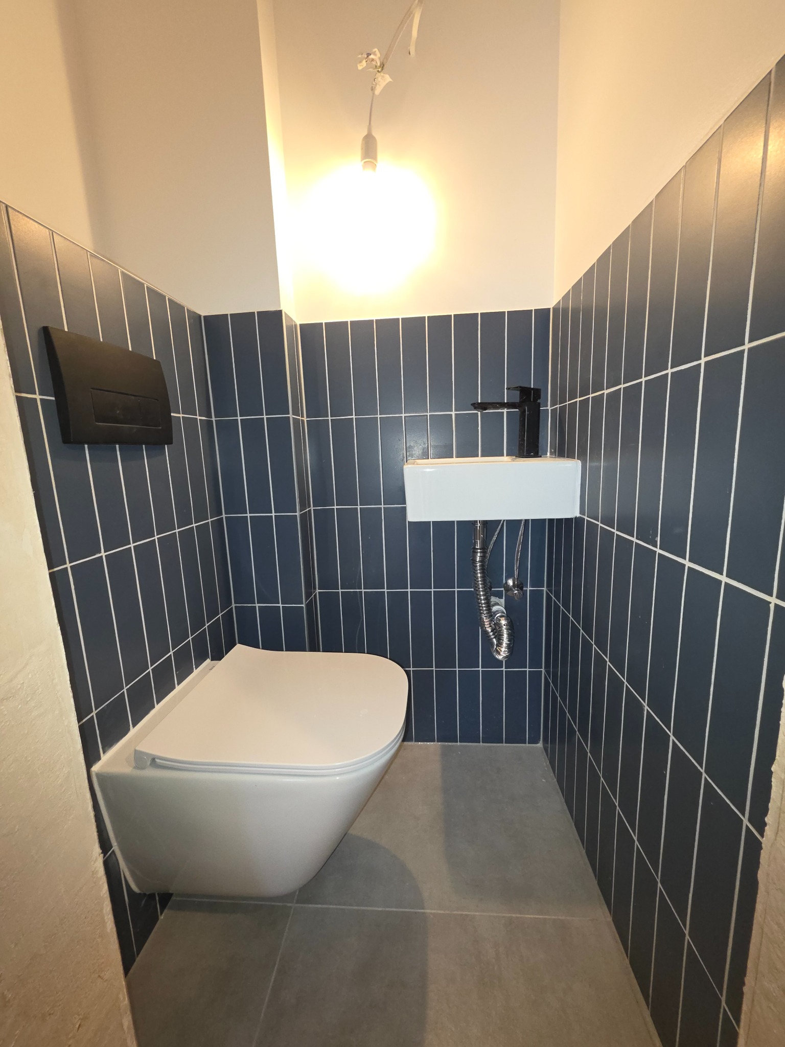 Kompaktowa łazienka z ciemnymi, pionowymi płytkami, podwieszaną toaletą i umywalką. Minimalistyczny design, czarna armatura, szara podłoga. Nowoczesne wykończenie.