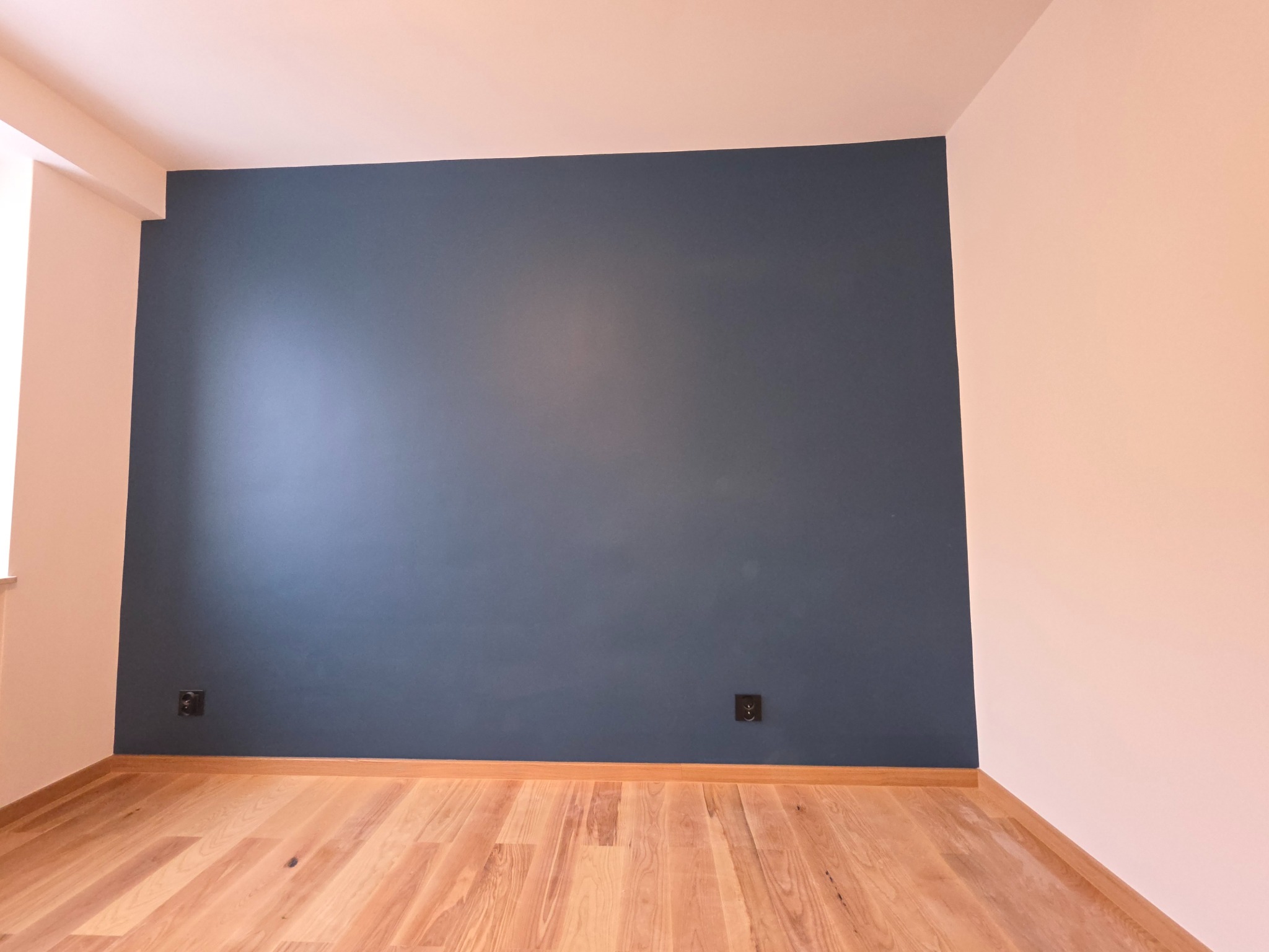 Pomalowane na granatowo ściany w pokoju z drewnianą podłogą. Widoczne gniazdka elektryczne i fragment okna z lewej strony. Ściany w odcieniach różu.
