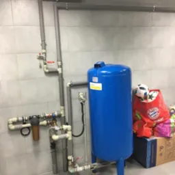 Niebieski hydrofor z podłączonymi rurami i filtrem wody, zamontowany na tle szarych płytek, z piłką i torbą na zakupy obok.