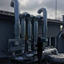 IGLO Mieczysław Klank - Montaż systemu wentylacji mechanicznej z rekuperacją na dachu budynku, widoczne rury wentylacyjne, centrala rekuperacyjna, pracownik w czarnym ubraniu oraz narzędzia pomiarowe.