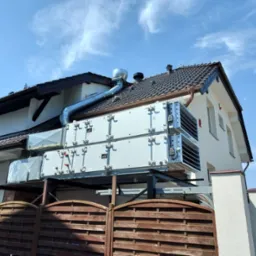 Dwa moduły rekuperacji VTS zamontowane na zewnątrz budynku jednorodzinnego, z widocznymi kanałami wentylacyjnymi i brązowym ogrodzeniem w tle.