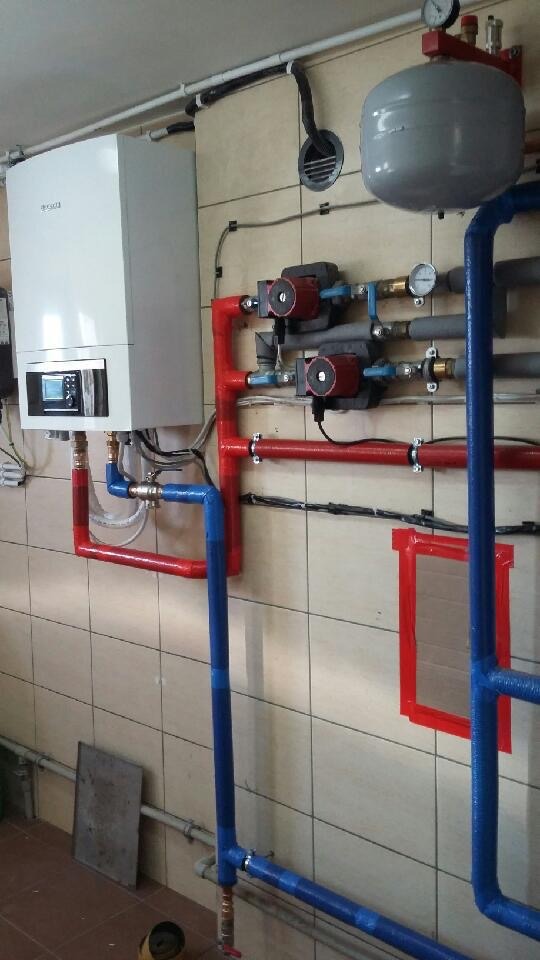 Biały piec gazowy kondensacyjny z podłączonymi czerwonymi i niebieskimi rurami, widoczne pompy obiegowe i zbiornik wyrównawczy, zamontowany na ścianie z kwadratowych płytek.