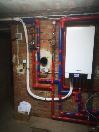 Montaż pieca gazowego w Pucku z widocznymi rurami w kolorze czerwonym i niebieskim, zamontowanego na ceglanej ścianie. Na podłodze widoczny karton z dokumentami.