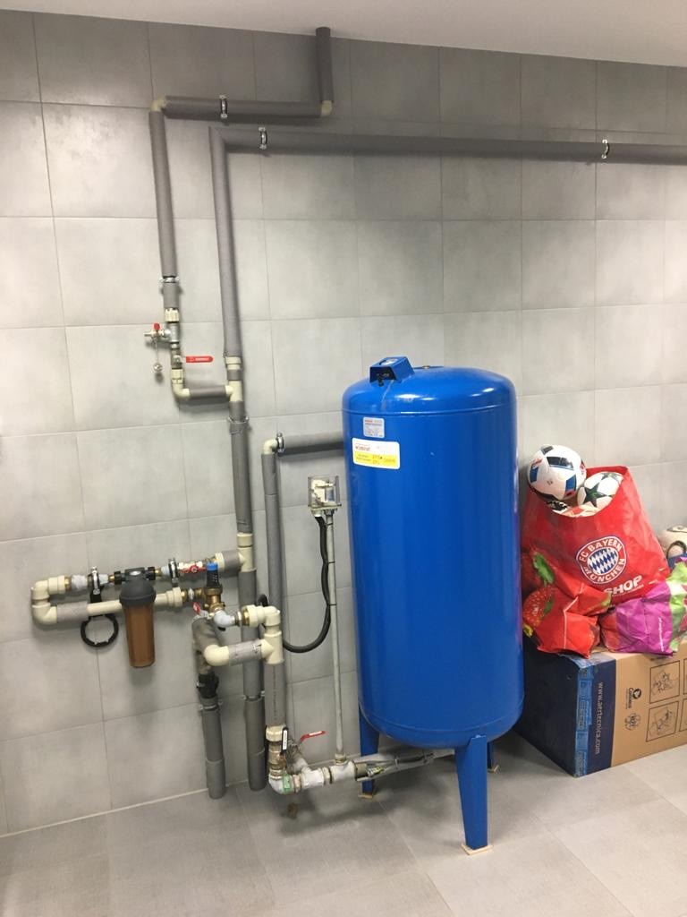 Niebieski hydrofor z podłączonymi rurami i filtrem wody, zamontowany na tle szarych płytek, z piłką i torbą na zakupy obok.