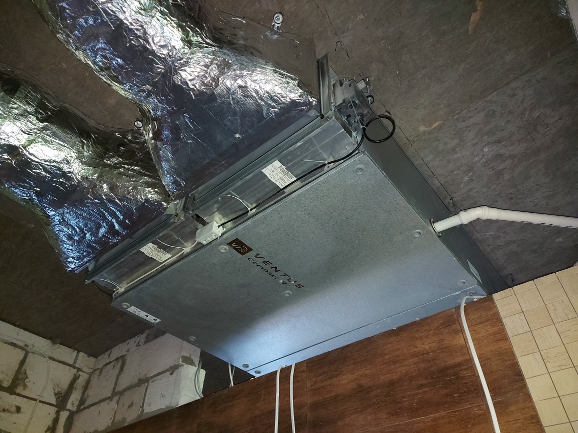 Zamontowana jednostka rekuperacyjna VTS Ventus Compact pod sufitem, widoczne rury wentylacyjne w izolacji aluminiowej, przewody elektryczne oraz fragment ściany z bloczków i paneli.
