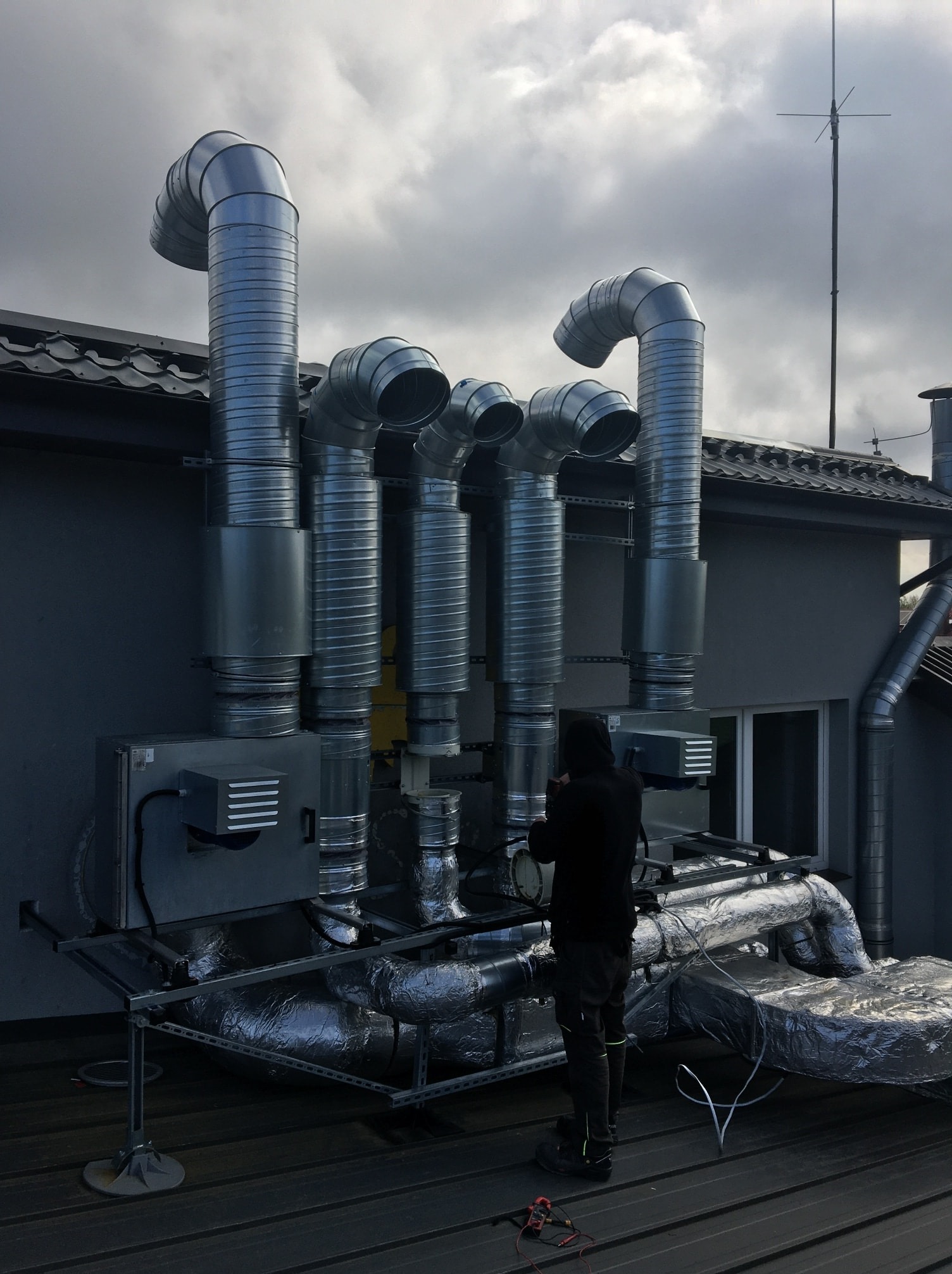 Montaż systemu wentylacji mechanicznej z rekuperacją na dachu budynku, widoczne rury wentylacyjne, centrala rekuperacyjna, pracownik w czarnym ubraniu oraz narzędzia pomiarowe.