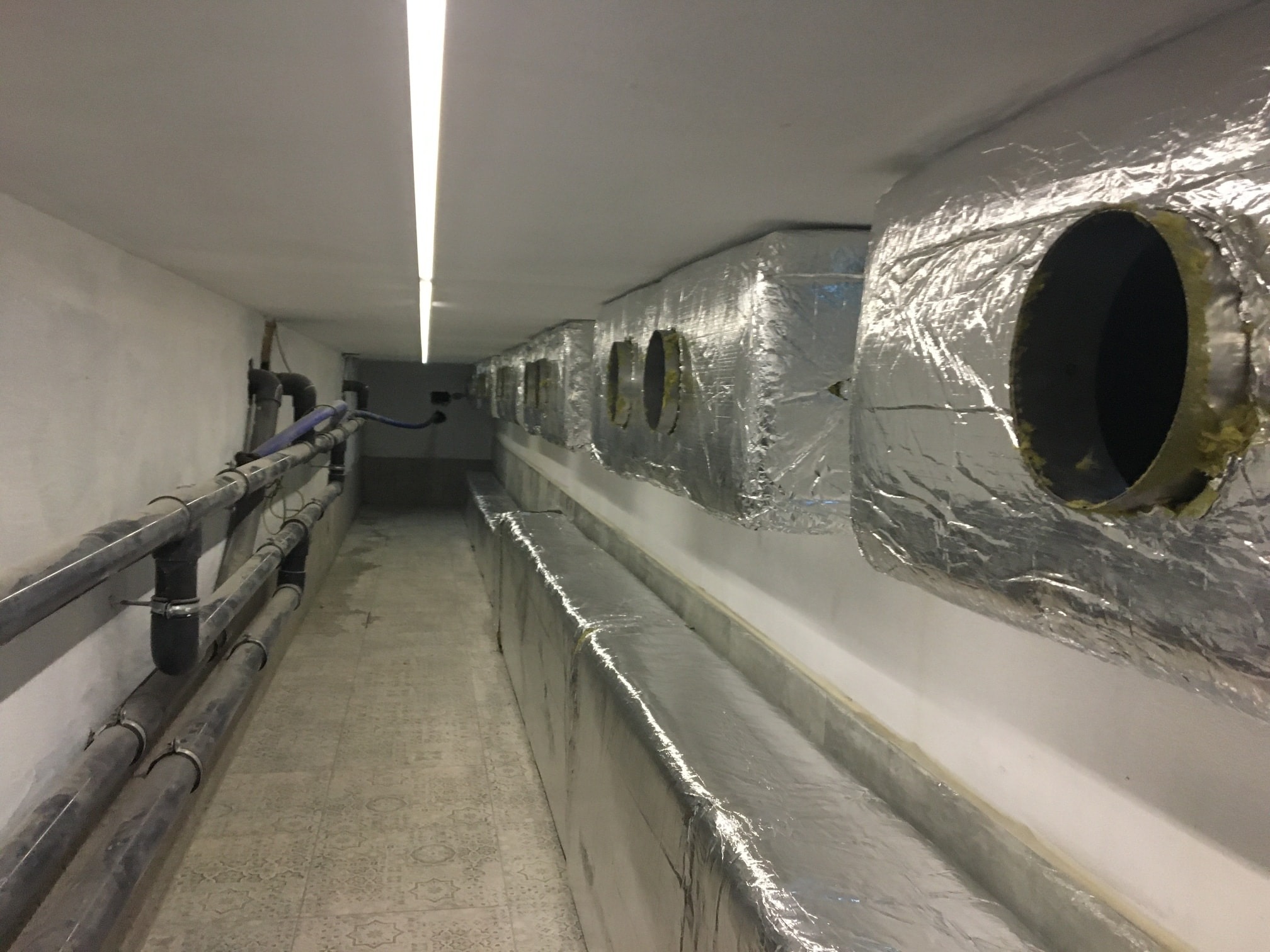 Korytarz techniczny z rzędami izolowanych przewodów wentylacyjnych i rur, oświetlony pojedynczą lampą LED na suficie.