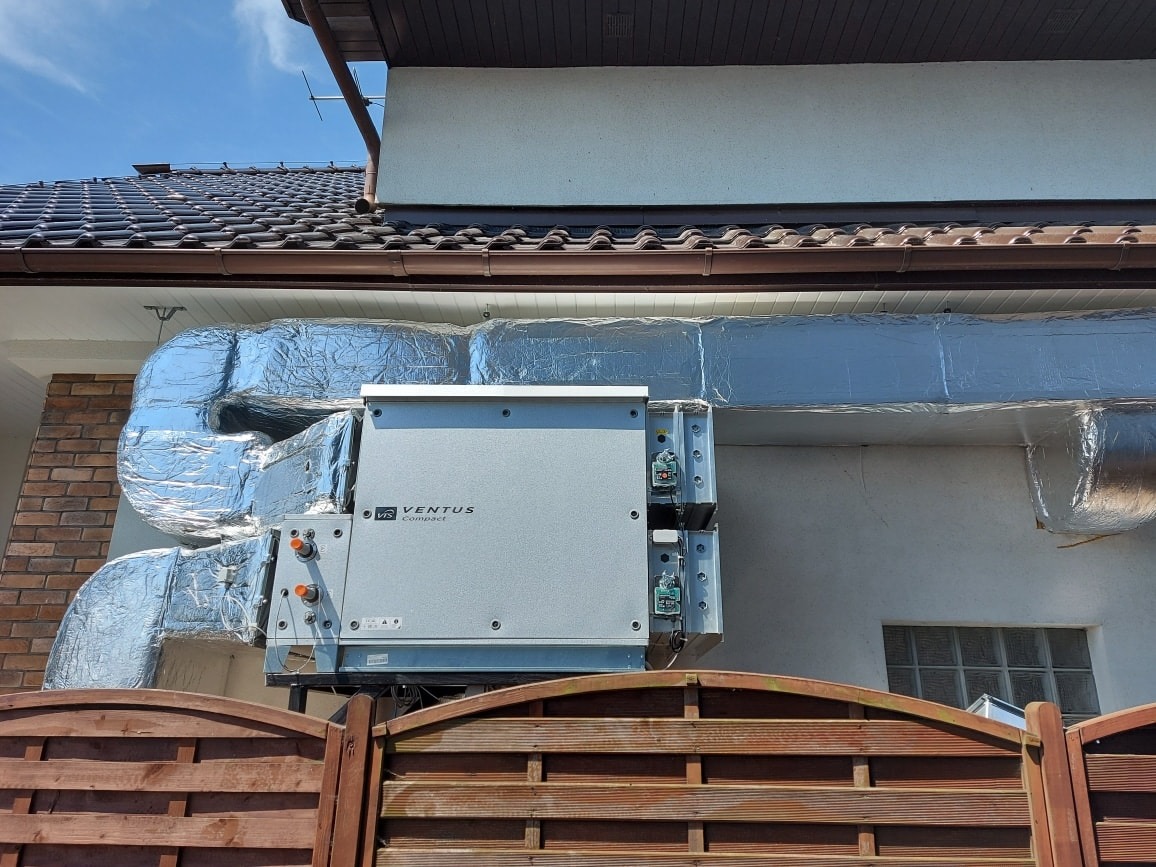 Szary rekuperator Ventus Compact zamontowany na zewnątrz budynku jednorodzinnego, z widocznymi przewodami izolowanymi folią aluminiową, ujęcie zza drewnianego płotu.