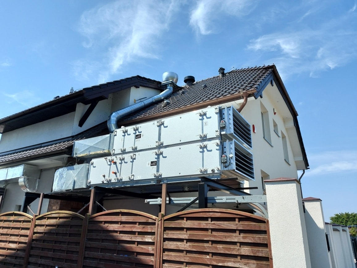 Dwa moduły rekuperacji VTS zamontowane na zewnątrz budynku jednorodzinnego, z widocznymi kanałami wentylacyjnymi i brązowym ogrodzeniem w tle.