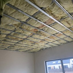 STORO-BUD Oleksandr Storozhenko - Montaż izolacji z wełny mineralnej w metalowym stelażu sufitu podwieszanego w jasnym pomieszczeniu z oknem. Widoczna część drabiny i fragment sylwetki montera.