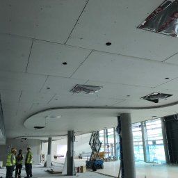 STORO-BUD Oleksandr Storozhenko - Montaż płyt gipsowo-kartonowych na suficie w nowoczesnym biurze. Widoczne otwory na oświetlenie i wentylację, pracownicy w odblaskowych kamizelkach i podnośnik.