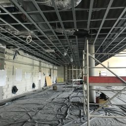 STORO-BUD Oleksandr Storozhenko - Sufit podwieszany w trakcie montażu w dużej przestrzeni biurowej. Widoczne rusztowania, instalacje i zabezpieczenia podłogi folią. Surowy stan ścian.