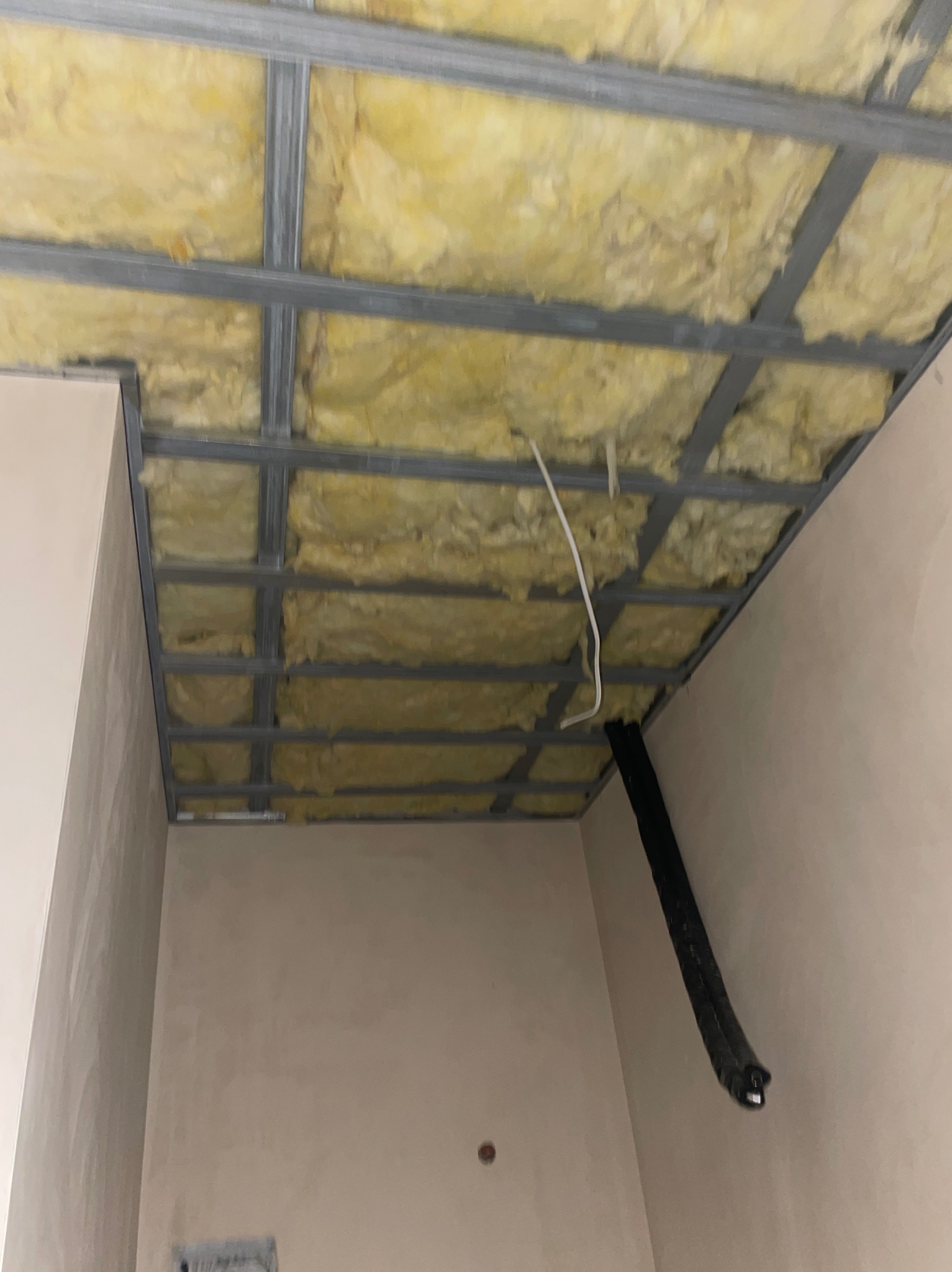 Sufit podwieszany z izolacją z wełny mineralnej, widoczne profile metalowe i fragment ściany w jasnym odcieniu. Ujęcie z dołu, perspektywa zbieżna.