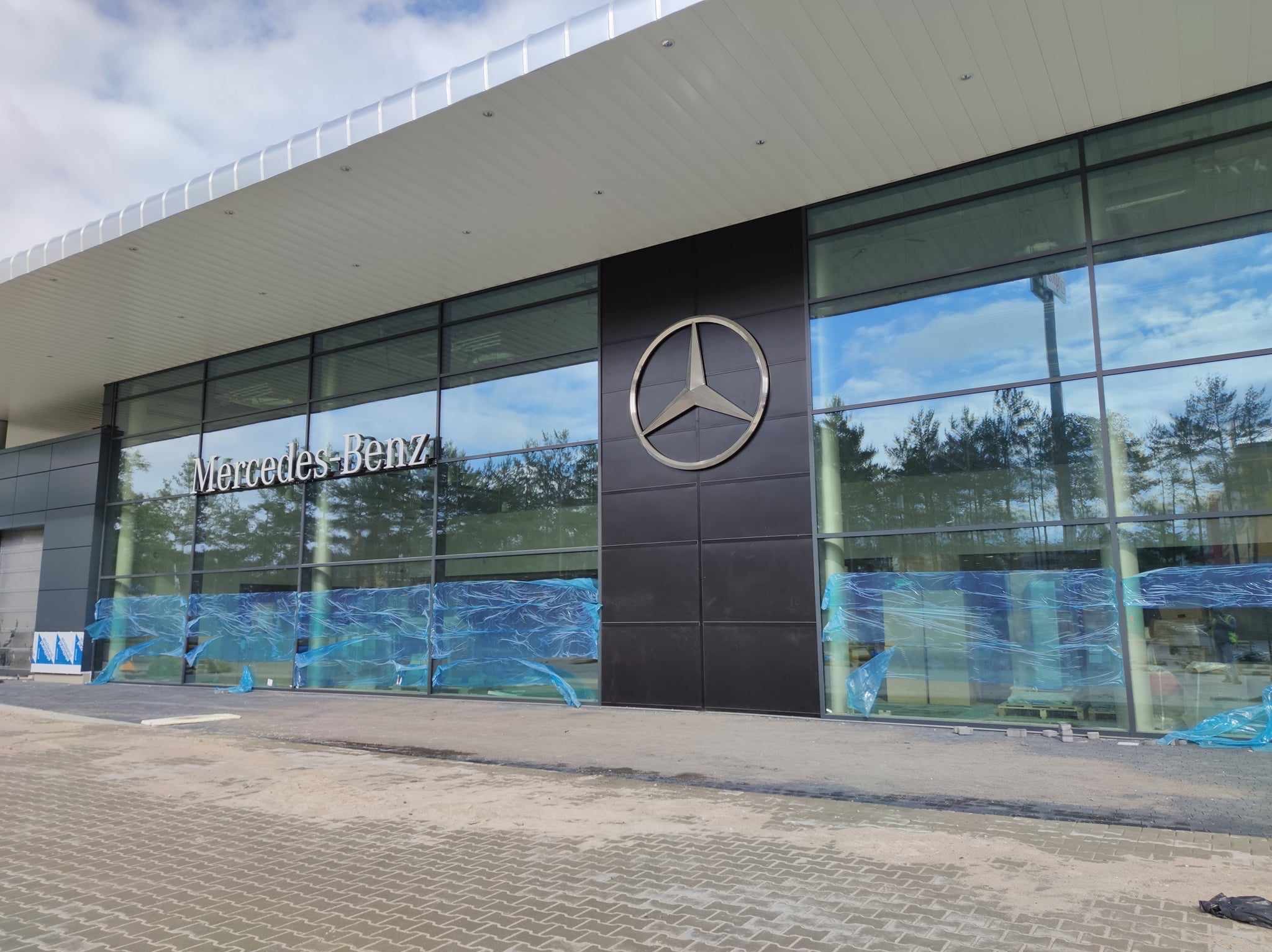 Nowoczesny salon Mercedes-Benz z przeszkloną fasadą, logo na czarnym panelu i zabezpieczeniami z folii na oknach, sugerującymi prace wykończeniowe.