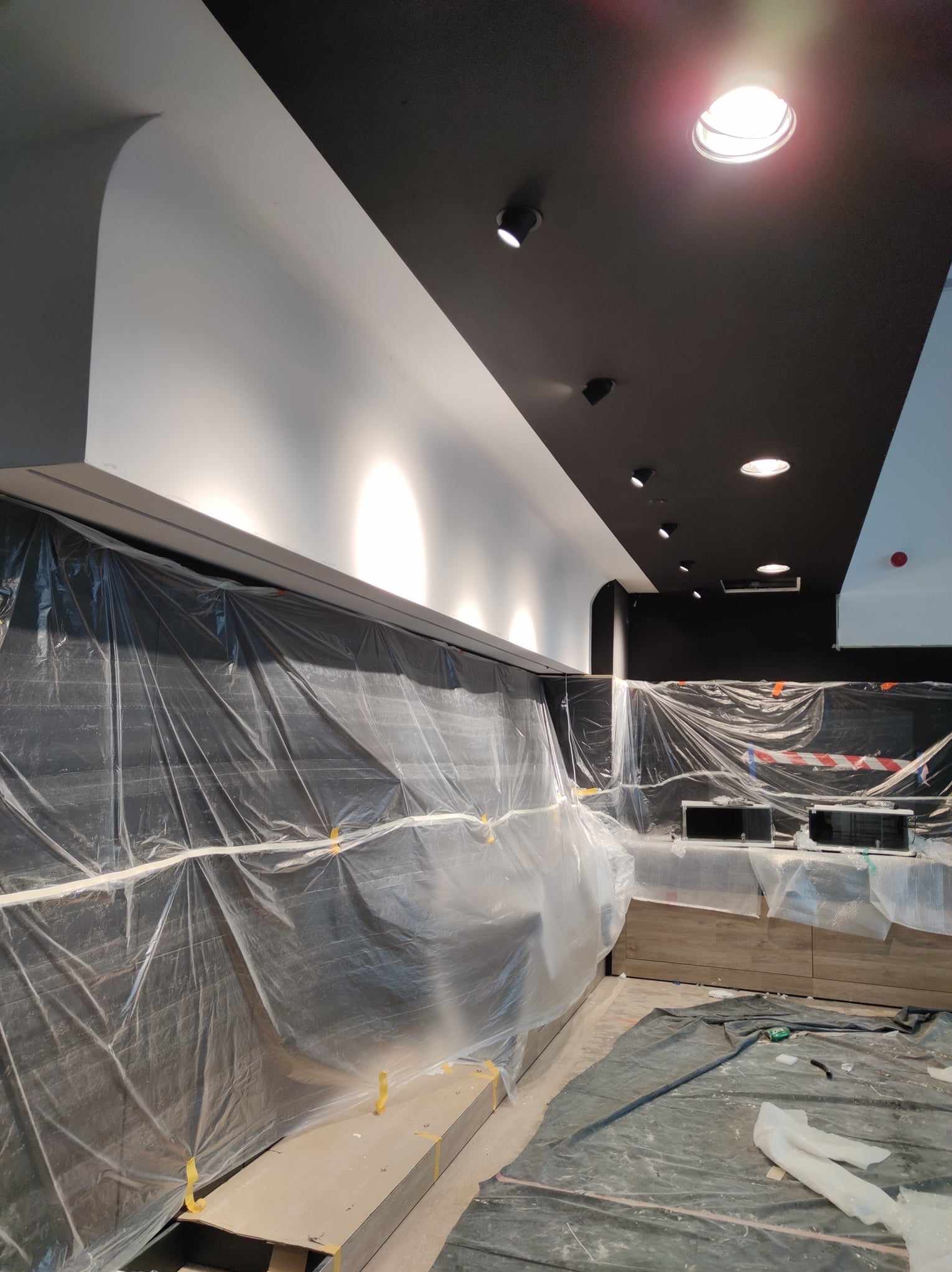 Ciemny sufit z oświetleniem punktowym i białe ściany w trakcie prac wykończeniowych, zabezpieczone folią malarską. Widoczne elementy instalacji.