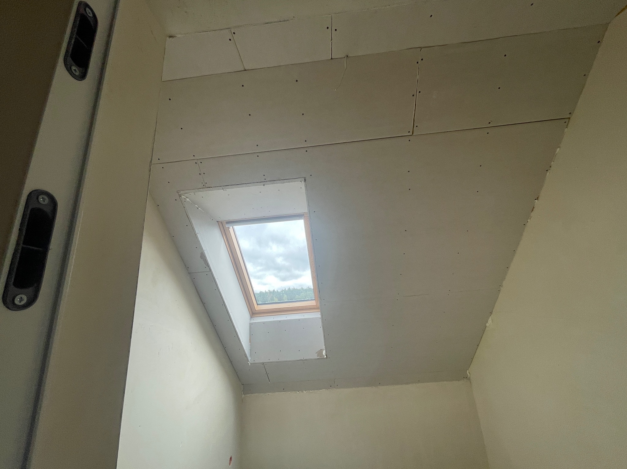 Montaż płyt karton-gips na poddaszu z oknem dachowym. Widoczne łączenia płyt i wkręty. Poziomica przy ścianie. Wnętrze w trakcie prac wykończeniowych.