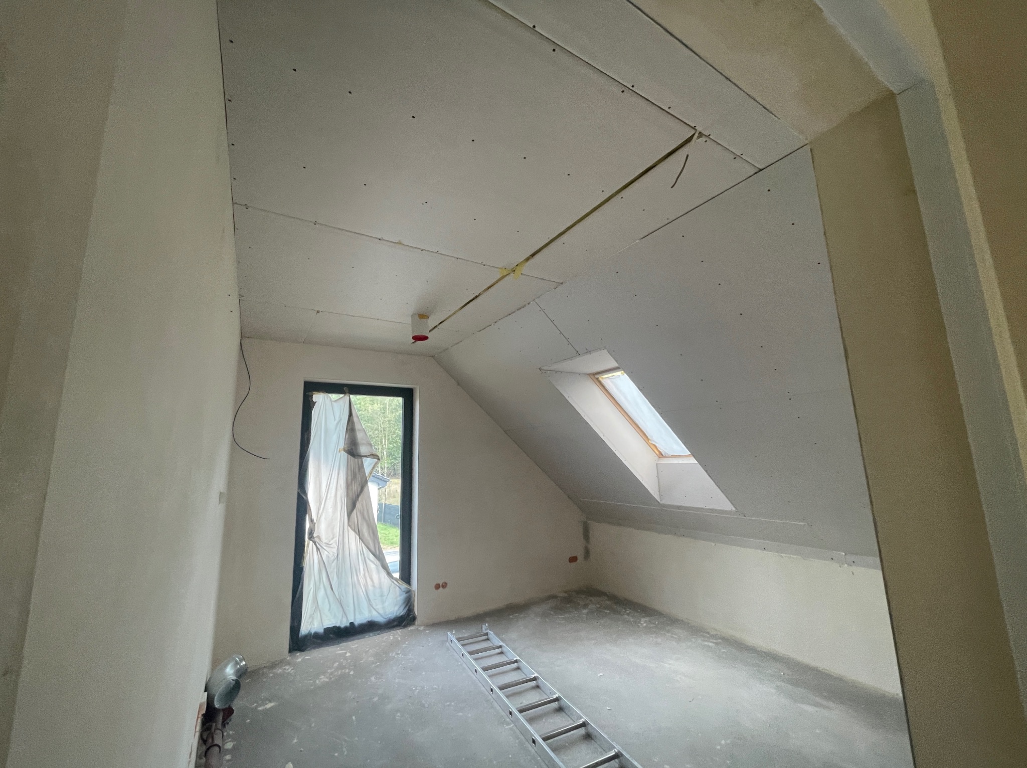 Pokój na poddaszu w trakcie montażu płyt kartonowo-gipsowych na ścianach i suficie. Widoczne okno dachowe, drabina i folia zabezpieczająca okno.