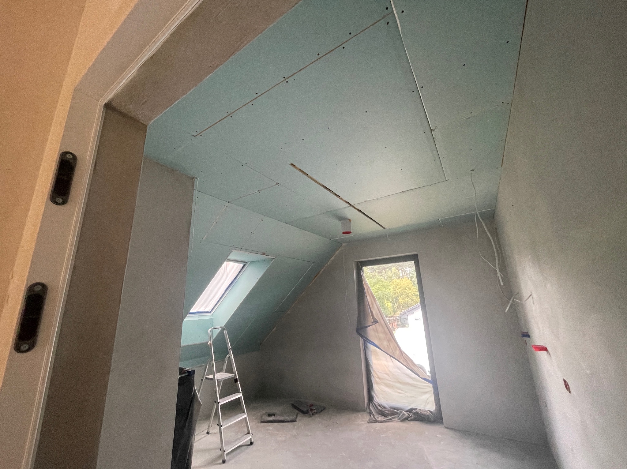 Poddasze w trakcie remontu: zamontowana płyta karton-gips na skośnym suficie, ściany w szpachli, drabina oparta o ścianę, okno dachowe, zabezpieczone drzwi.