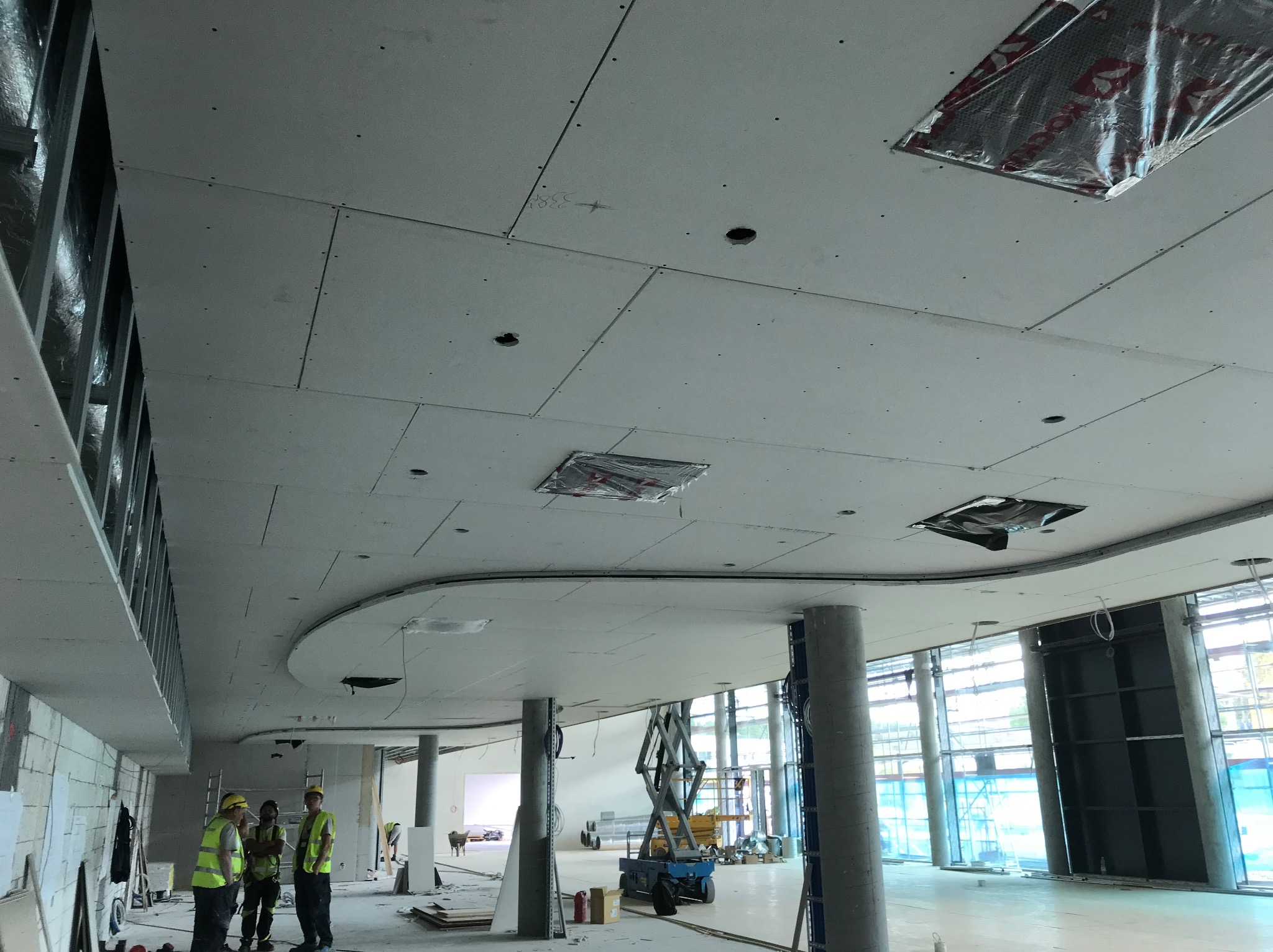 Montaż płyt gipsowo-kartonowych na suficie w nowoczesnym biurze. Widoczne otwory na oświetlenie i wentylację, pracownicy w odblaskowych kamizelkach i podnośnik.