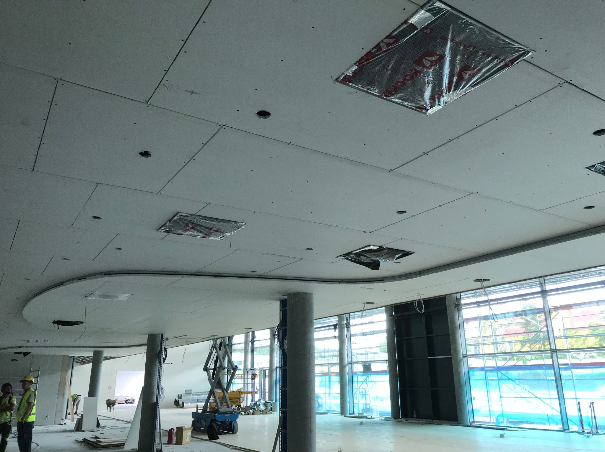 Montaż sufitu podwieszanego z płyt kartonowo-gipsowych w dużej przestrzeni biurowej. Widoczne otwory na oświetlenie i elementy instalacji. Prace w toku, widoczny podnośnik.