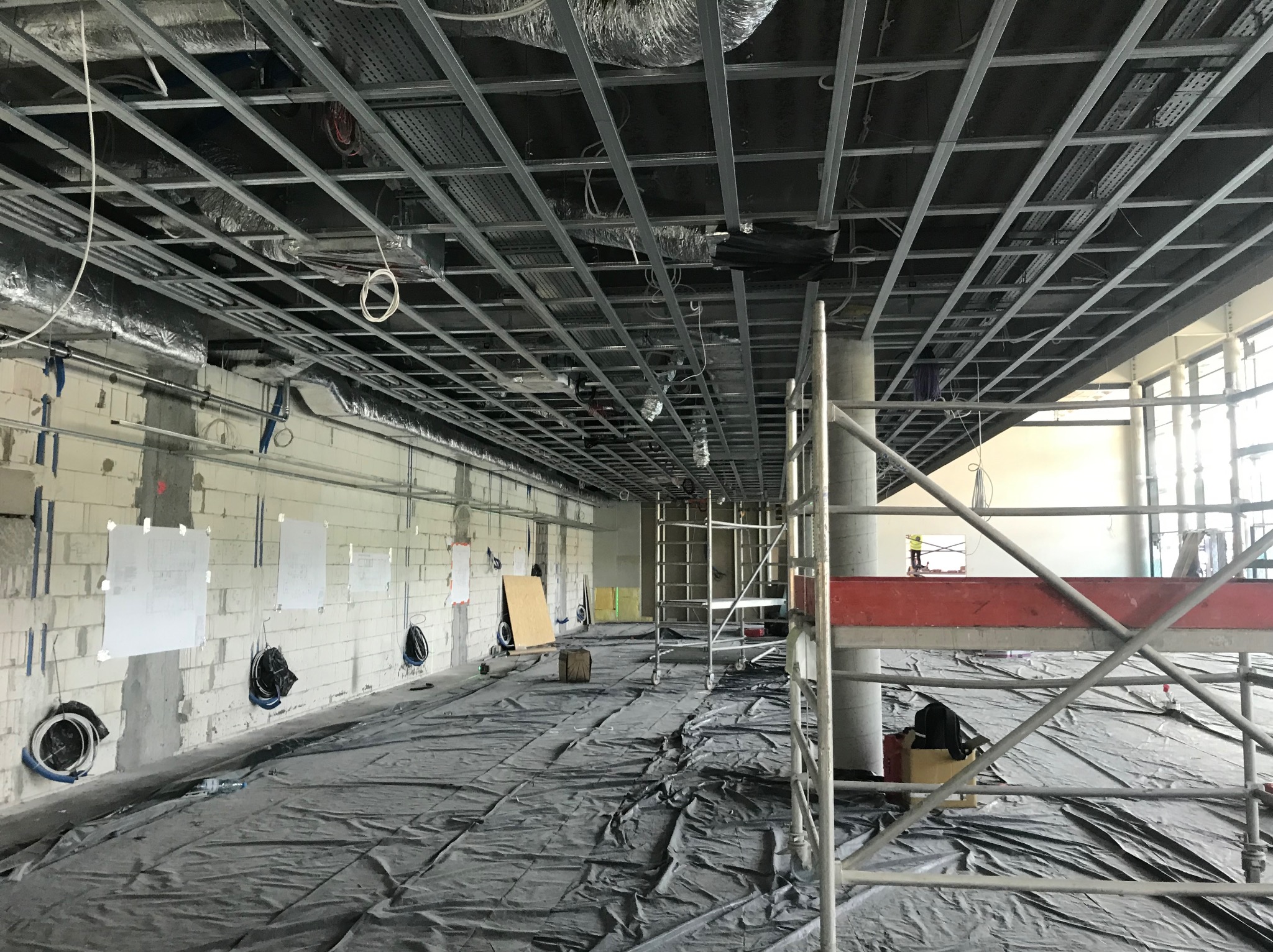 Sufit podwieszany w trakcie montażu w dużej przestrzeni biurowej. Widoczne rusztowania, instalacje i zabezpieczenia podłogi folią. Surowy stan ścian.