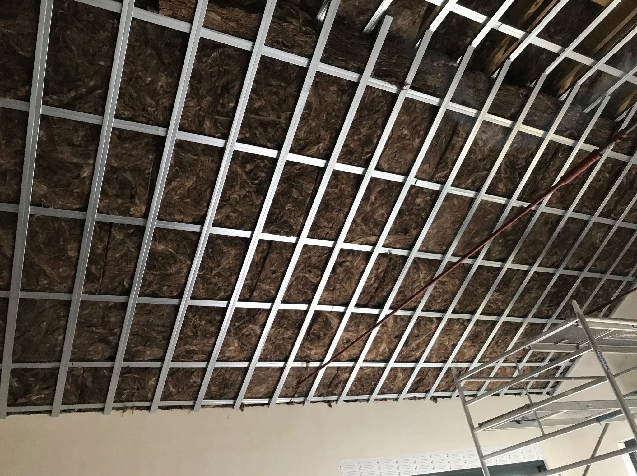 Metalowa konstrukcja sufitu podwieszanego z izolacją z wełny mineralnej, widok z dołu, fragment rusztowania w prawym dolnym rogu.