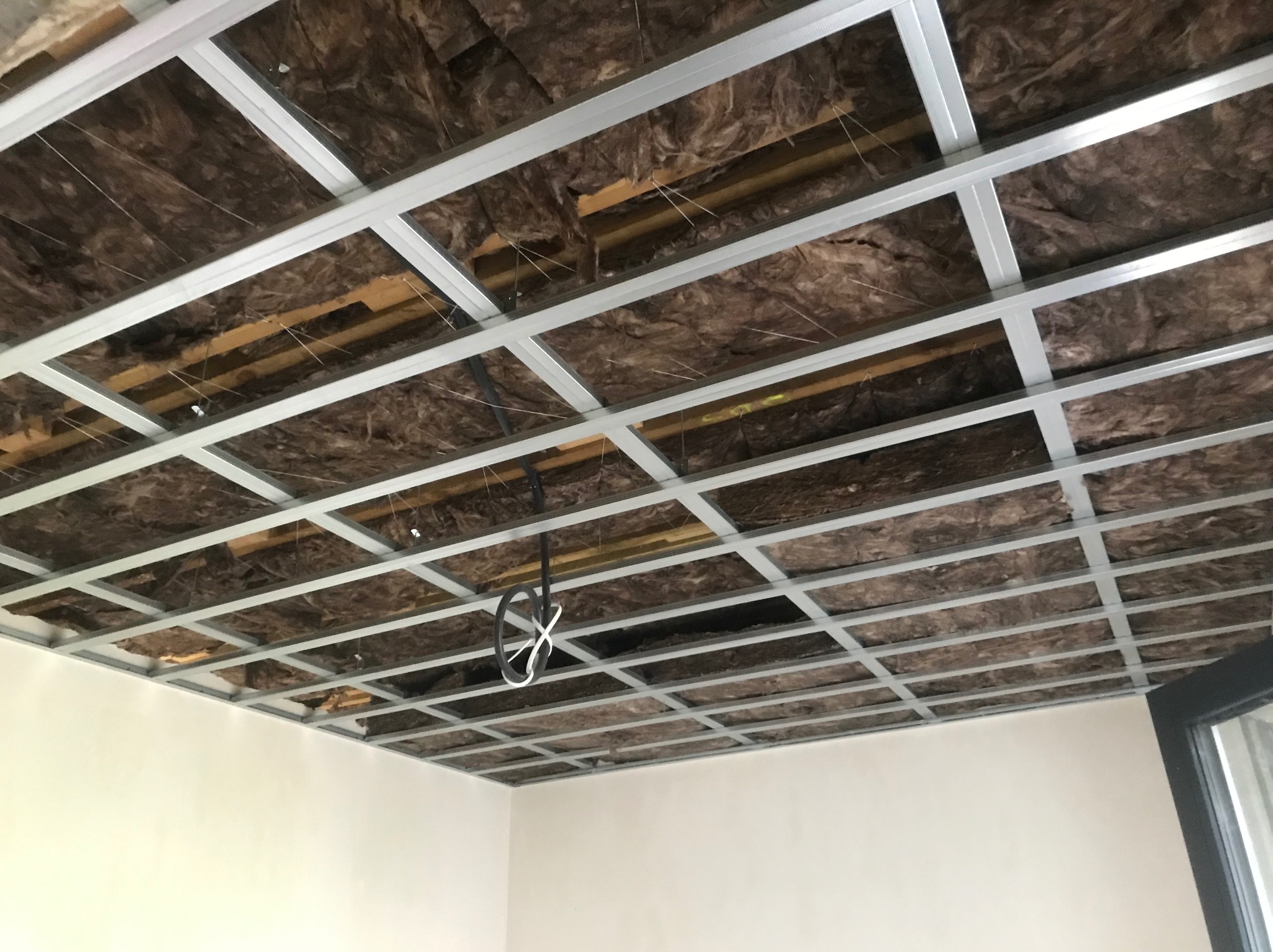 Montaż sufitu podwieszanego z widoczną izolacją z wełny mineralnej i metalową konstrukcją. Widok z dołu na częściowo ukończony sufit.