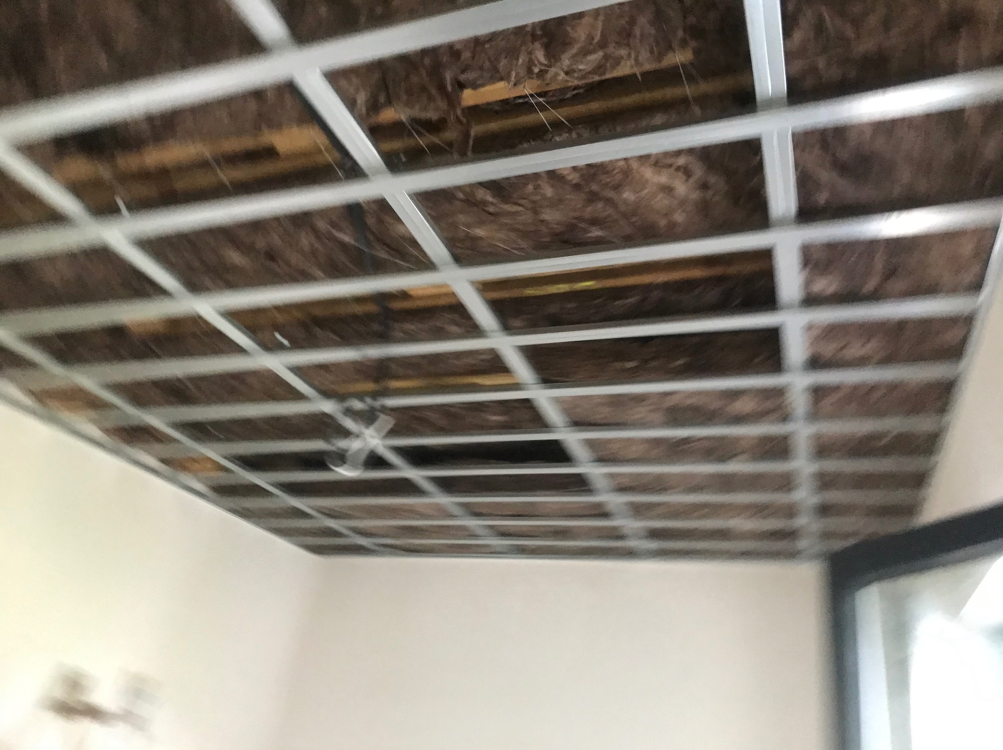 Szkielet sufitu podwieszanego z widoczną izolacją, metalowa konstrukcja w kwadratowej siatce, widok z dołu, fragment okna po prawej.