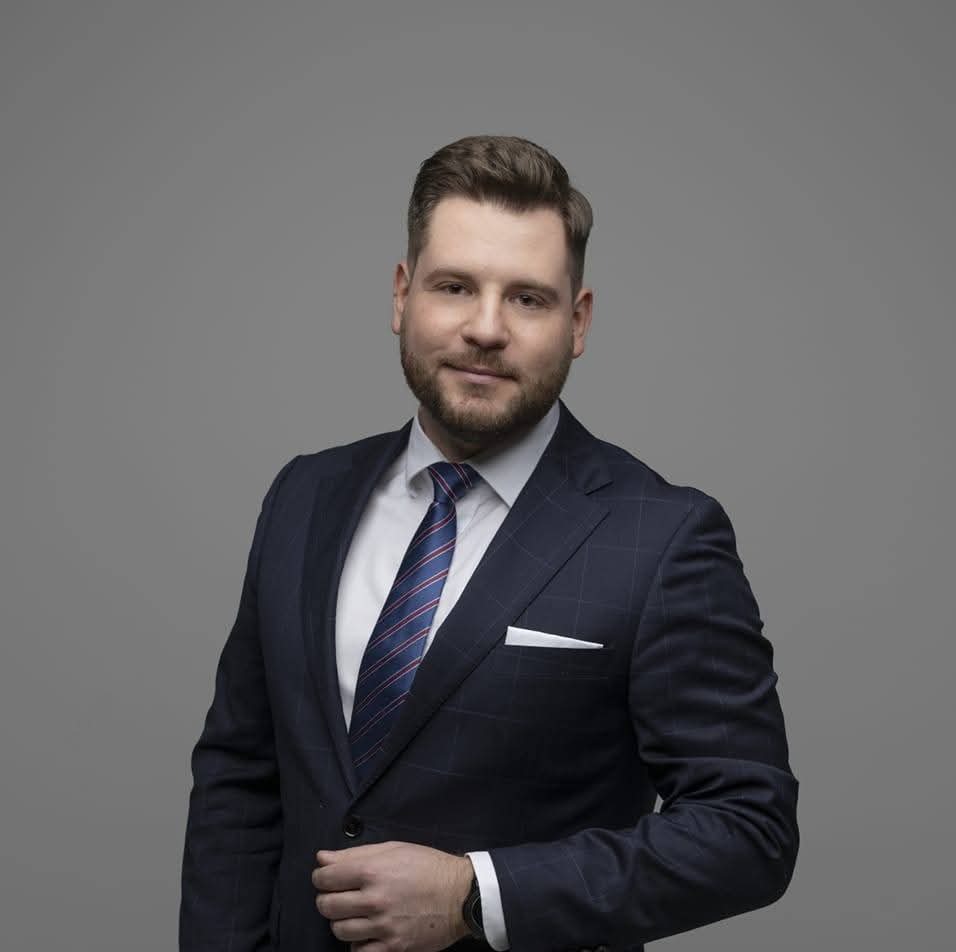 Mężczyzna w granatowym garniturze w kratę i krawacie w paski, z białą poszetką w kieszeni, na szarym tle. Uśmiechnięty, z brodą, patrzy w obiektyw.