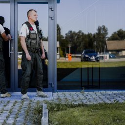 Kontakt Ochrona Sp. z o.o. - Dwóch ochroniarzy w mundurach i kamizelkach taktycznych stoi przed nowoczesnym budynkiem z przeszklonymi drzwiami, w których odbija się zaparkowany samochód i otoczenie.