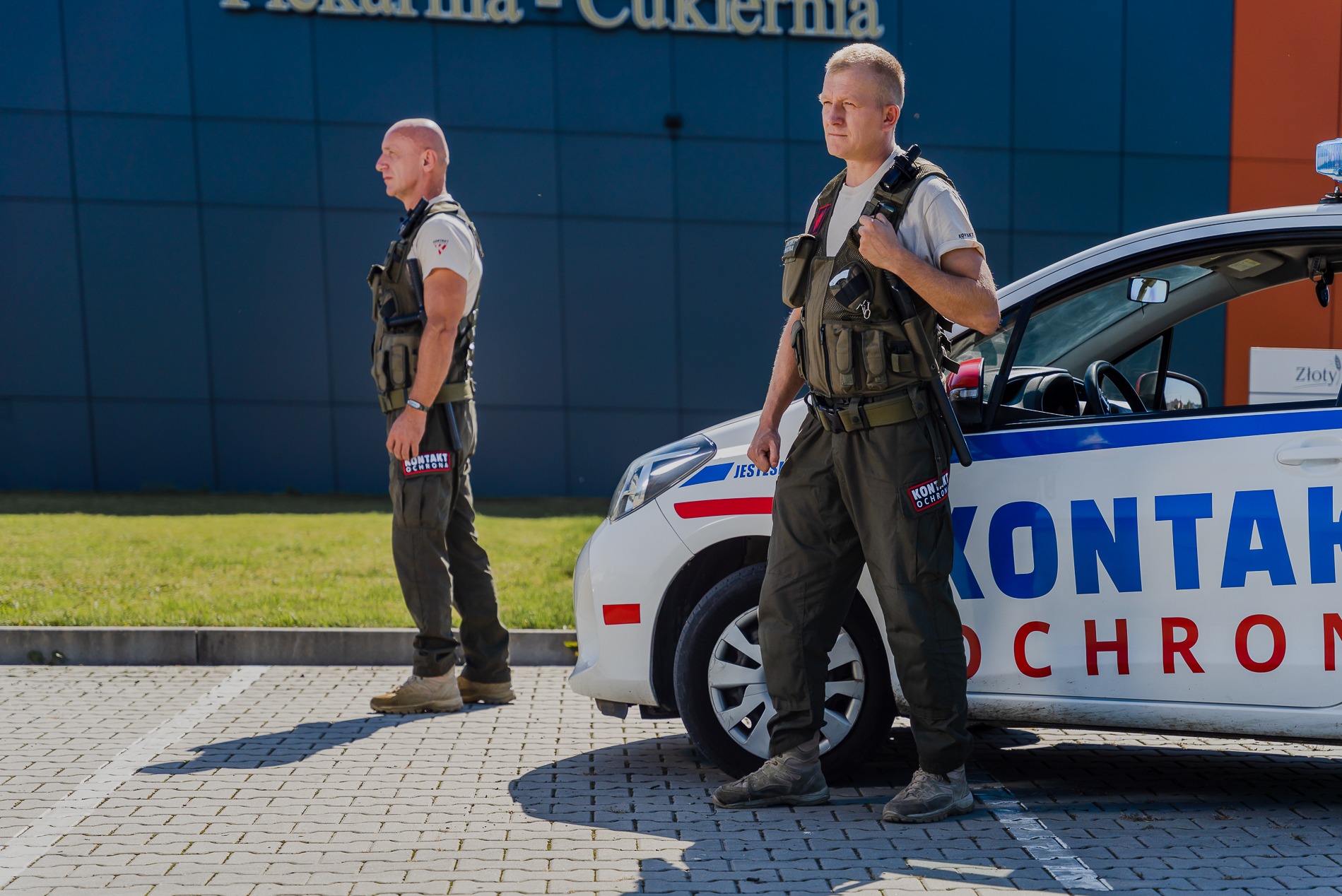 Dwóch ochroniarzy w kamizelkach taktycznych stoi obok oznakowanego samochodu patrolowego przed budynkiem z napisem 'Piekarnia-Cukiernia'.