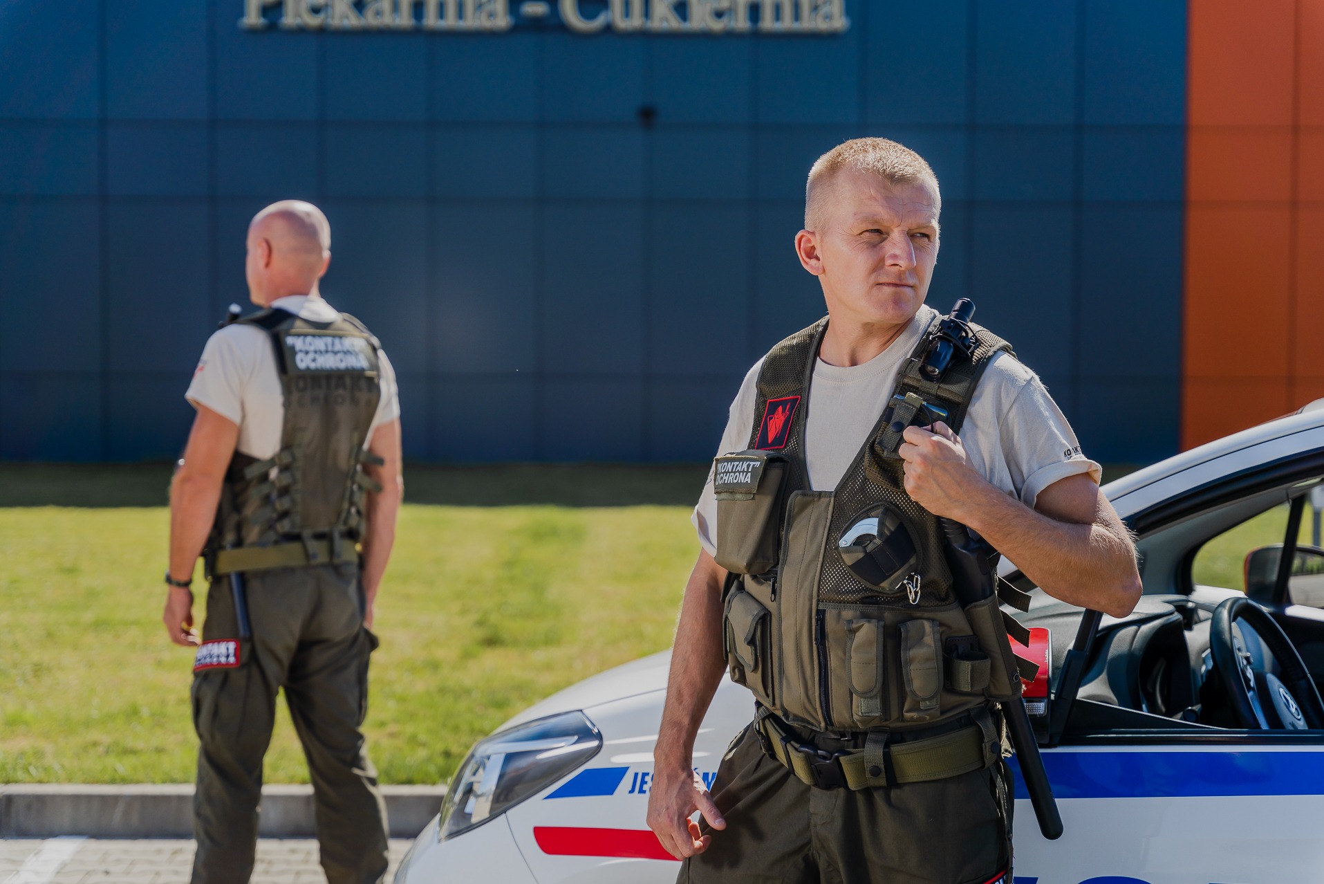 Dwóch ochroniarzy w kamizelkach taktycznych przed budynkiem piekarni-cukierni i samochodem patrolowym.